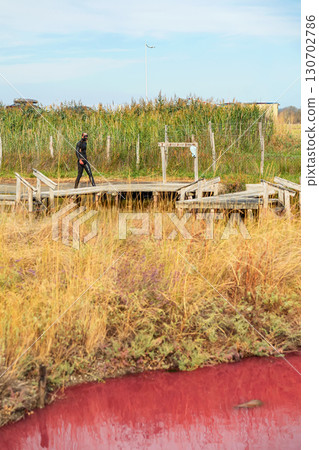 Man in black mud walks across wooden boardwalk near therapeutic lake surrounded by tall grass. Concept of mud spa, health escape, natural detox experience 130702786