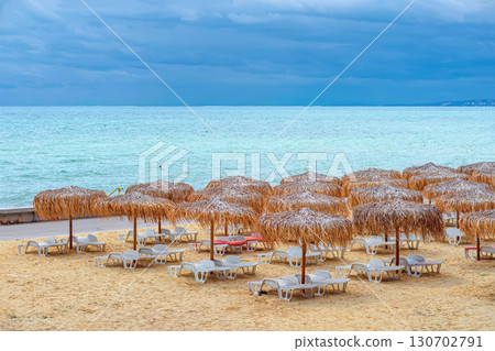 Empty sun loungers and straw umbrellas on sandy beach under moody sky, signaling end of summer season. Concept of autumn beach, off-season travel, vacation nostalgia Empty sun loungers and straw umbrellas on sandy beach under moody sky, signaling end of summer season. Concept of autumn beach, off-season travel, vacation nostalgia 130702791