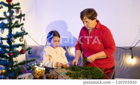 Grandmother and granddaughter decorate for Christmas, carefully arranging festive greenery together. 130703930