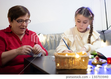 A grandmother and granddaughter spend quality time together, working on a project at a table with fairy lights. 130703940