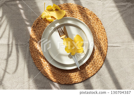 Table Setting with Yellow Leaves on White Plate and Woven Placemat under Sun Light Table Setting with Yellow Leaves on White Plate and Woven Placemat under Sun Light 130704387