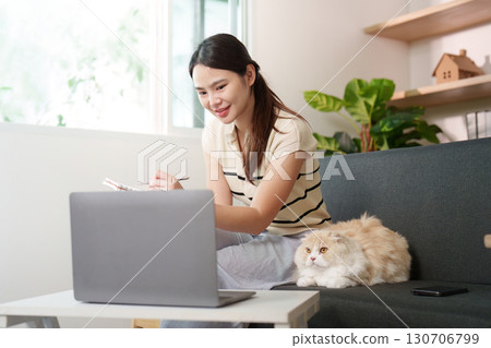 Productive Home Office. Young woman taking notes while working on laptop with cat nearby. 130706799