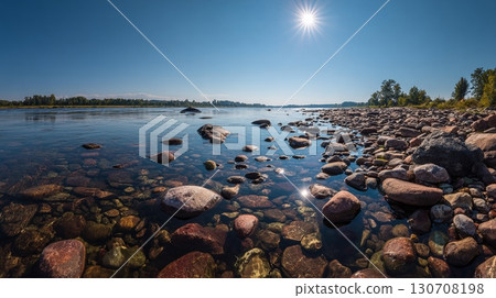 Clear water laps against a rocky shore under a brilliant sun, showcasing the serenity of nature's artistry and the beauty of a pristine river landscape. 130708198