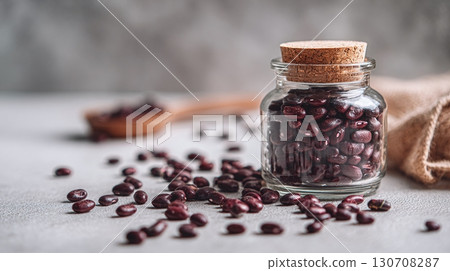 Beans in glass jar. The jar is sealed with a cork stopper, and beans are scattered around it on a light surface. There's also a wooden spoon with beans. 130708287