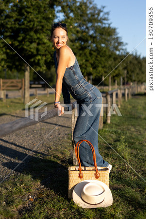 Young adult beautiful woman wear stylish jeans one-shoulder denim jumpsuit have fun walks outdoors warm sunny day holding straw bag hat hand. Female person carefree smiles enjoying countryside nature 130709356
