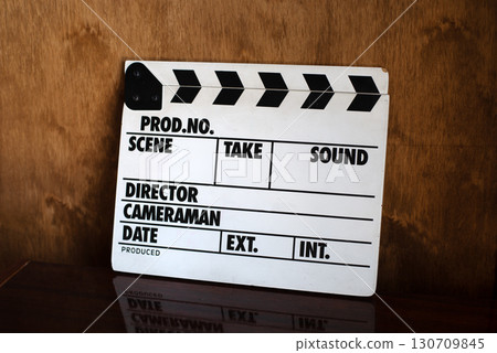 Classic film clapperboard resting on a dark, reflective surface against a warm wooden background.  130709845