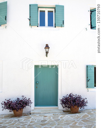 Turquoise wooden door with shutters on white plastered wall, purple potted plants, stone pavement 130710760