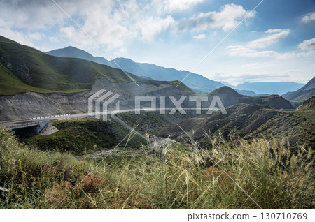 Mountainous terrain with deep valleys, green slopes and winding road under cloudy blue sky. 130710769