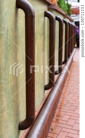 Brown metal railing with curved metal supports against a textured green wall, creating a rustic atmosphere in an outdoor setting with brick pathway 130711151