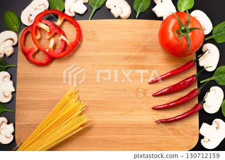A cutting board, raw spaghetti, fresh tomato, chili peppers, and mushrooms, framed on a dark background. 130712159