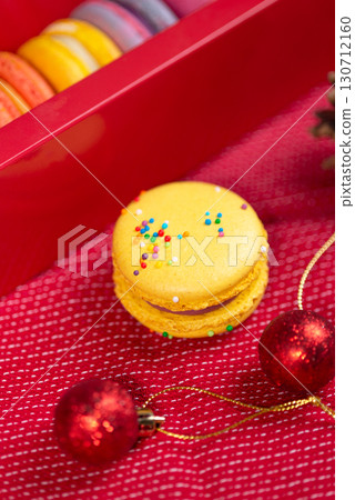 A bright yellow macaron with sprinkles, alongside red Christmas ornaments and a gift box, on a festive red background A bright yellow macaron with sprinkles, alongside red Christmas ornaments and a gift box, on a festive red background 130712160
