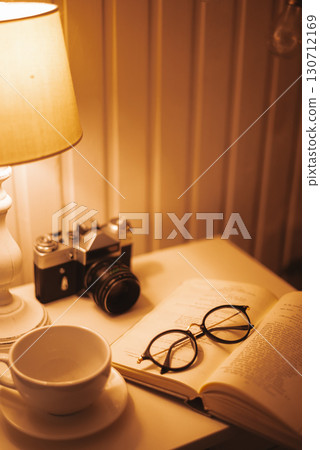 A cozy nightstand with an open book, reading glasses, a vintage camera, and a cup, all bathed in warm 130712169