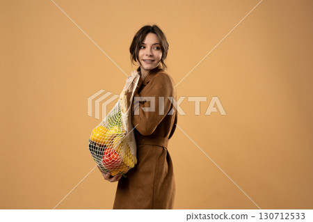 Young woman in brown coat holding eco mesh bag with vegetables on beige background, sustainable lifestyle and eco-friendly shopping concept 130712533