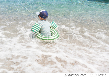 Rear view of a child sitting in a striped inflatable ring in shallow sea waves. Family-friendly beach moment suitable for parenting, safety campaigns, travel and childrens product adverts 130712954