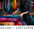 A skilled tailor arranges colorful fabrics in a shop, focused on her task while wearing a black tailor apron. The vibrant background features a variety of textiles showcasing her craftsmanship. 130712958