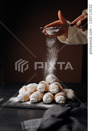 Fresh cream horns being dusted with powdered sugar on dark background, winter dessert vibes Fresh cream horns being dusted with powdered sugar on dark background, winter dessert vibes 130713032