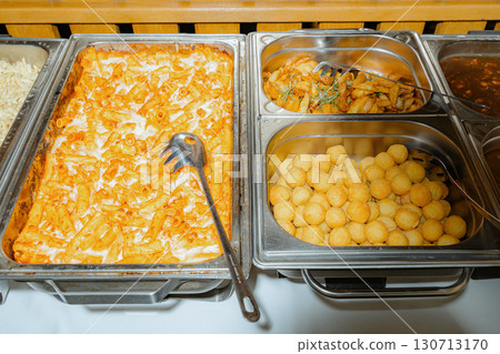 Buffet setup featuring baked pasta, crispy potatoes, and fried snacks in serving trays. Perfect for events, displaying a tempting variety of dishes for guests to enjoy Buffet setup featuring baked pasta, crispy potatoes, and fried snacks in serving trays. Perfect for events, displaying a tempting variety of dishes for guests to enjoy 130713170