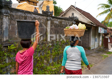 Local women carry baskets of food on their heads in an Indonesian village. Portrait of a smiling woman in Bali. 130713307