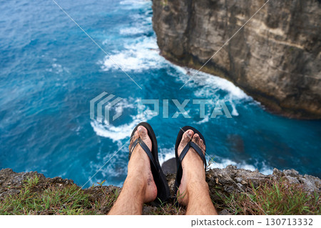 A man sits on a cliff with his legs dangling over an abyss above the ocean. First person photo. A man sits on a cliff with his legs dangling over an abyss above the ocean. First person photo. 130713332