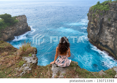 A beautiful woman in a pink dress sits on a cliff above the ocean on the island of Nusa Penida. Devil's Billabong an incredibly wonderful lagoon with splashes from the waves. 130713393