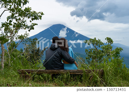 A couple of lovers are sitting on a bench, hugging each other and enjoying the view of the popular sacred cloud-covered Mount Agung on the island of Bali. A couple of lovers are sitting on a bench, hugging each other and enjoying the view of the popular sacred cloud-covered Mount Agung on the island of Bali. 130713431