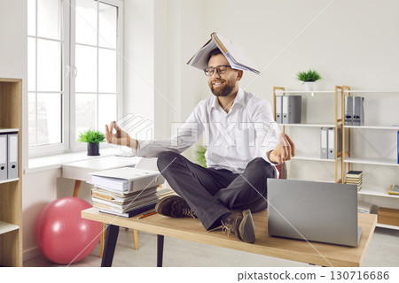 Funny business man with book on head sitting on office desk, doing yoga and meditating 130716686