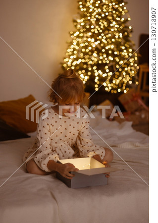 Girl opening glowing gift box sitting on bed near Christmas tree with lights in cozy room Girl opening glowing gift box sitting on bed near Christmas tree with lights in cozy room 130718907
