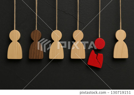 Wooden hanging figurines in various shades with one broken red piece symbolizing individuality and disruption against textured black background creating minimalist design 130719151