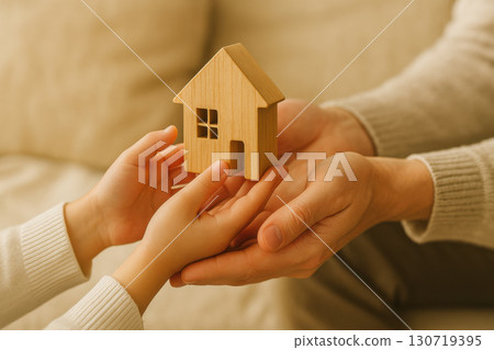 Wooden house figure held by child and adult hands symbolizing generational inheritance and real estate transfer with cozy warm background and soft lighting 130719395