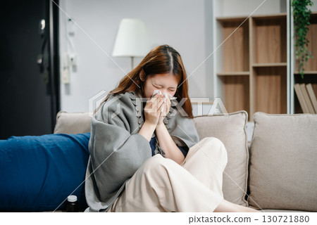 Young Asian woman suffering from flu symptoms, covered with a blanket on a sofa. Concept of illness, healthcare, and recovery. Young Asian woman suffering from flu symptoms, covered with a blanket on a sofa. Concept of illness, healthcare, and recovery. 130721880