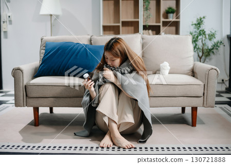 Young Asian woman suffering from flu symptoms, covered with a blanket on a sofa. Concept of illness, healthcare, and recovery. Young Asian woman suffering from flu symptoms, covered with a blanket on a sofa. Concept of illness, healthcare, and recovery. 130721888