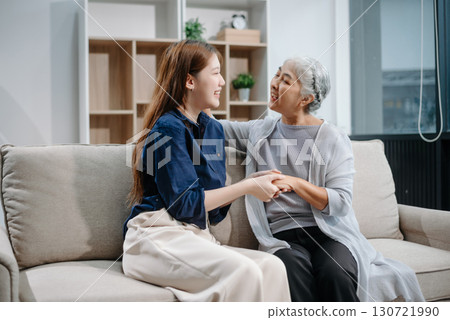 Happy adult Asian granddaughter and senior grandmother having fun enjoying talk sit on sofa in living room using a laptop, tablet 130721990