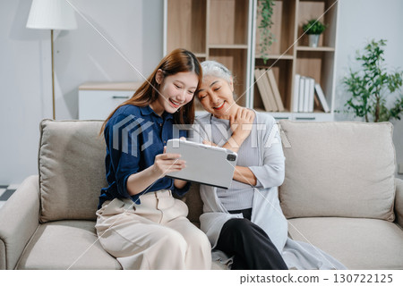 Happy adult Asian granddaughter and senior grandmother having fun enjoying talk sit on sofa in living room using a laptop, tablet Happy adult Asian granddaughter and senior grandmother having fun enjoying talk sit on sofa in living room using a laptop, tablet 130722125