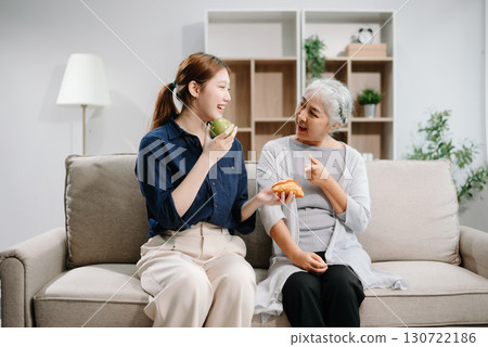 Happy adult Asian granddaughter and senior grandmother having fun enjoying talk sit on sofa in modern living room, using a laptop, tablet 130722186