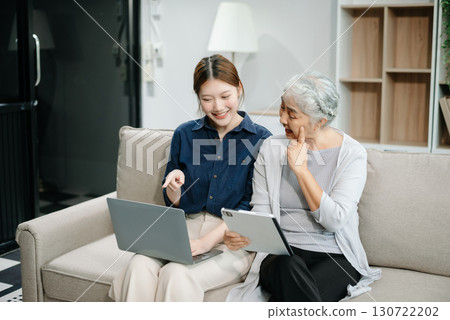 Happy adult Asian granddaughter and senior grandmother having fun enjoying talk sit on sofa in modern living room, using a laptop, tablet 130722202