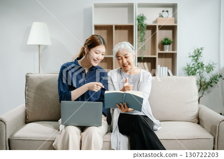 Happy adult Asian granddaughter and senior grandmother having fun enjoying talk sit on sofa in modern living room, using a laptop, tablet 130722203