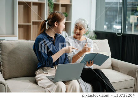 Happy adult Asian granddaughter and senior grandmother having fun enjoying talk sit on sofa in modern living room, using a laptop, tablet 130722204