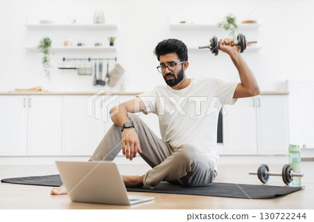 Young adult man sits on exercise mat in home kitchen lifting dumbbells by laptop. Engages in remote fitness routine, demonstrating home workout. Young adult man sits on exercise mat in home kitchen lifting dumbbells by laptop. Engages in remote fitness routine, demonstrating home workout. 130722244