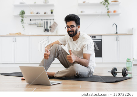 Young adult man recording video blog about sports and fitness at home, sitting on yoga mat, showing thumbs up to laptop. Capturing content on healthy lifestyle, indoors in home kitchen setting. Young adult man recording video blog about sports and fitness at home, sitting on yoga mat, showing thumbs up to laptop. Capturing content on healthy lifestyle, indoors in home kitchen setting. 130722263