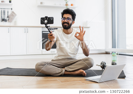Young adult man with black beard and glasses records sports vlog at home. Sits on yoga mat in modern interior, using smartphone and tripod, shows gesture, promotes health 130722266