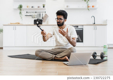 Young adult man recording video blog about fitness and health at home, sitting cross-legged on mat, using smartphone mounted on stick, with laptop and water bottle nearby in bright kitchen setting. 130722275