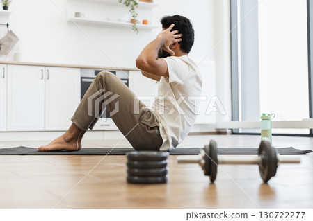 Young adult man exercises at home performing sit-ups on mat in kitchen setting promoting fitness, health commitment, and active lifestyle motivation. Young adult man exercises at home performing sit-ups on mat in kitchen setting promoting fitness, health commitment, and active lifestyle motivation. 130722277