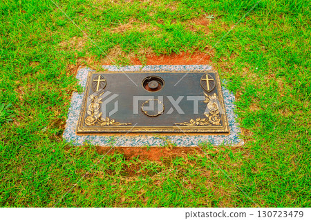 Gravestone featuring ornate carvings, symbols rests on lush green grass in calm cemetery environment. Gravestone featuring ornate carvings, symbols rests on lush green grass in calm cemetery environment. 130723479