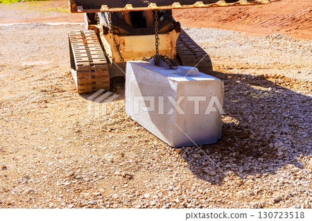 Construction vehicle carefully lifts places concrete block onto gravel surface under works day 130723518