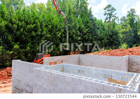 Workers are actively pouring concrete into constructed foundation walls of building amidst lush greenery. Workers are actively pouring concrete into constructed foundation walls of building amidst lush greenery. 130723544