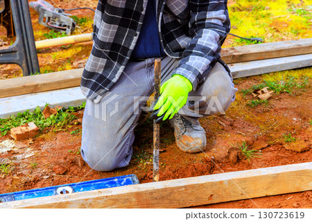 Worker uses tool to dig while kneeling on ground, ensuring proper alignment at construction site. Worker uses tool to dig while kneeling on ground, ensuring proper alignment at construction site. 130723619