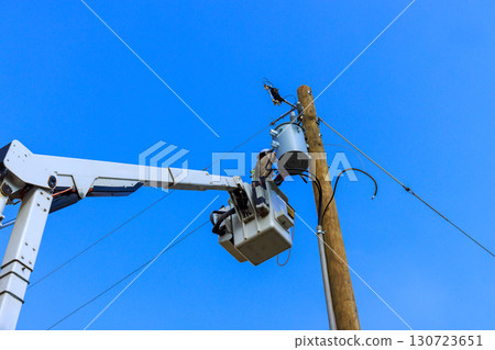 Utility electrical worker uses bucket truck to fix an electrical transformer atop wooden pole against 130723651