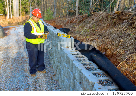 Construction worker wearing safety gear inspects stone retaining wall alignment while working on landscaping project. Construction worker wearing safety gear inspects stone retaining wall alignment while working on landscaping project. 130723700