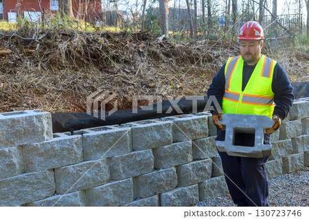 Construction worker in safety vest,helmet lifts block for retaining wall Construction worker in safety vest,helmet lifts block for retaining wall 130723746
