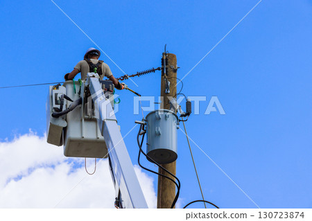 Utility worker inspects electrical equipment atop bucket truck under bright sunlight few clouds. 130723874